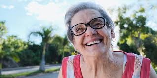 A man with medium-length wavy hair, wearing a teal work jacket, stands looking directly at the camera with a serious expression. The background features blurred white containers, suggesting a workplace environment.